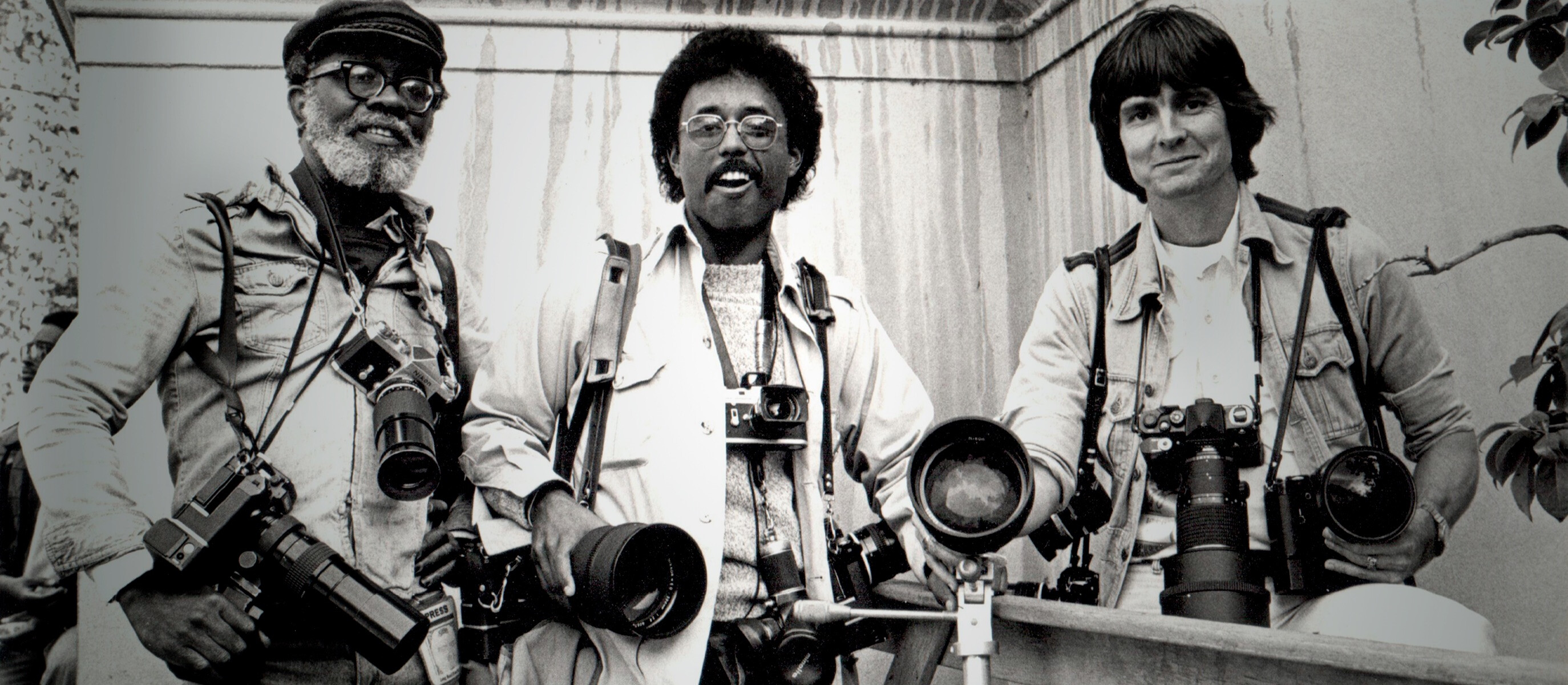 A black and white photo of photographer Veryl Oakland wearing several cameras around his neck, standing with two other photographers.